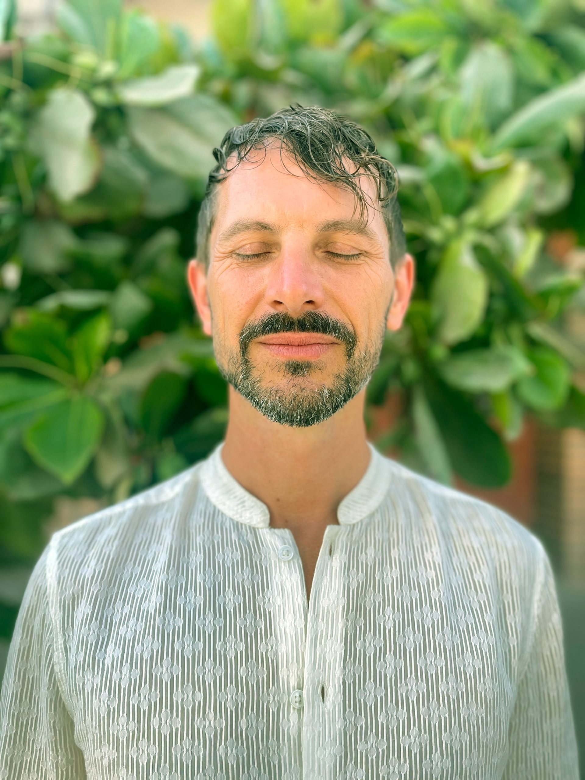 Smiling man with eyes closed in front of lush green leaves, embodying inner peace and grounded strength.